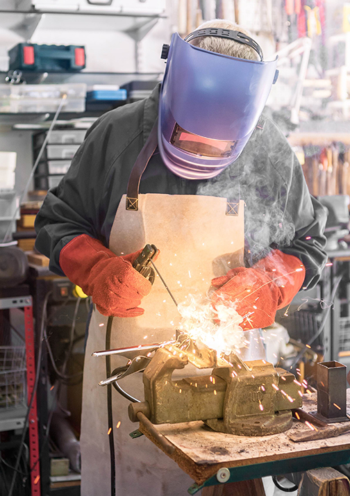 A man welder with safety helmet working with arc welding machine in the workshop, sideways flashes. Male hands in gloves fix a piece of iron with sparks fly in different directions.