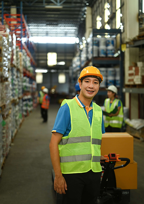 Male warehouse worker in hardhats and reflective jackets pulling a pallet truck walking through retail warehouse full of shelves.